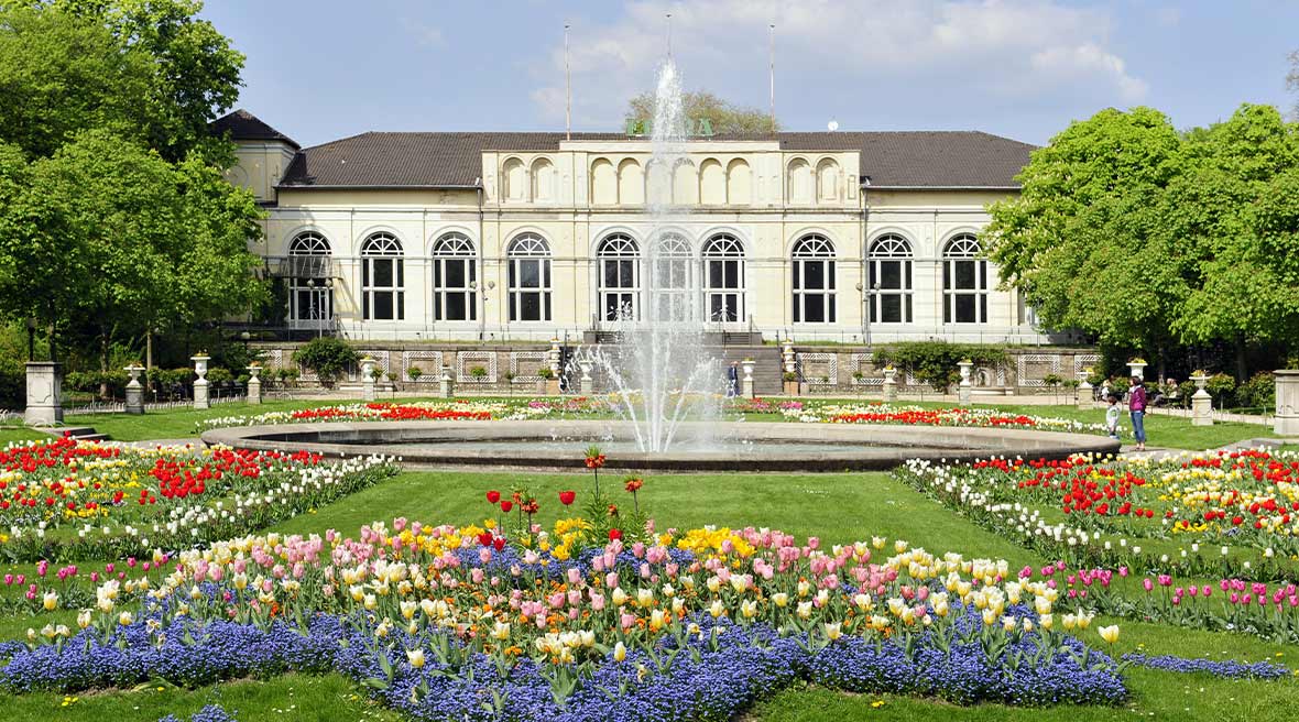 White building with green trees either side of it with green lawns and flower beds and fountain